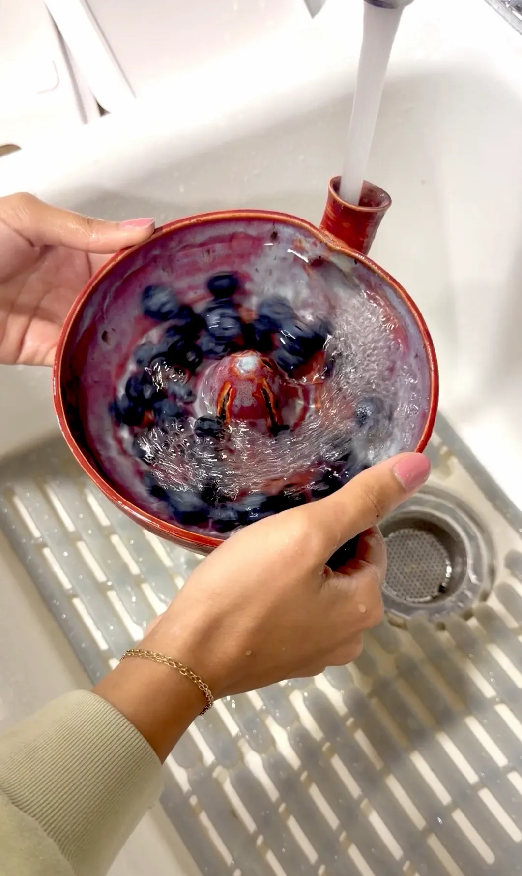 Hands holding a red berry bowl under a running tap, water swirling around blueberries.