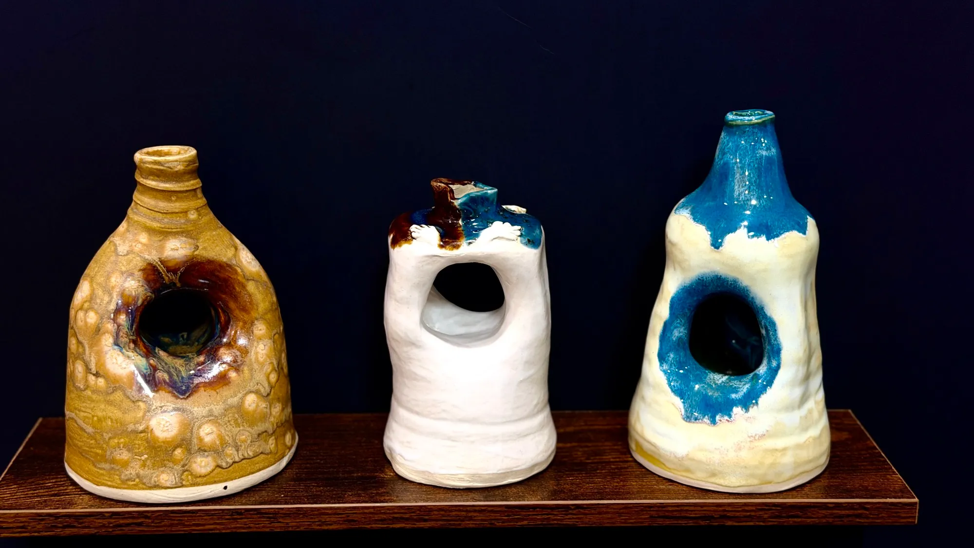 Three tall vessels on a wooden shelf, each with a hole through its body — one speckled gold, one white with a blue and brown top, one cream with a pooled blue glaze.