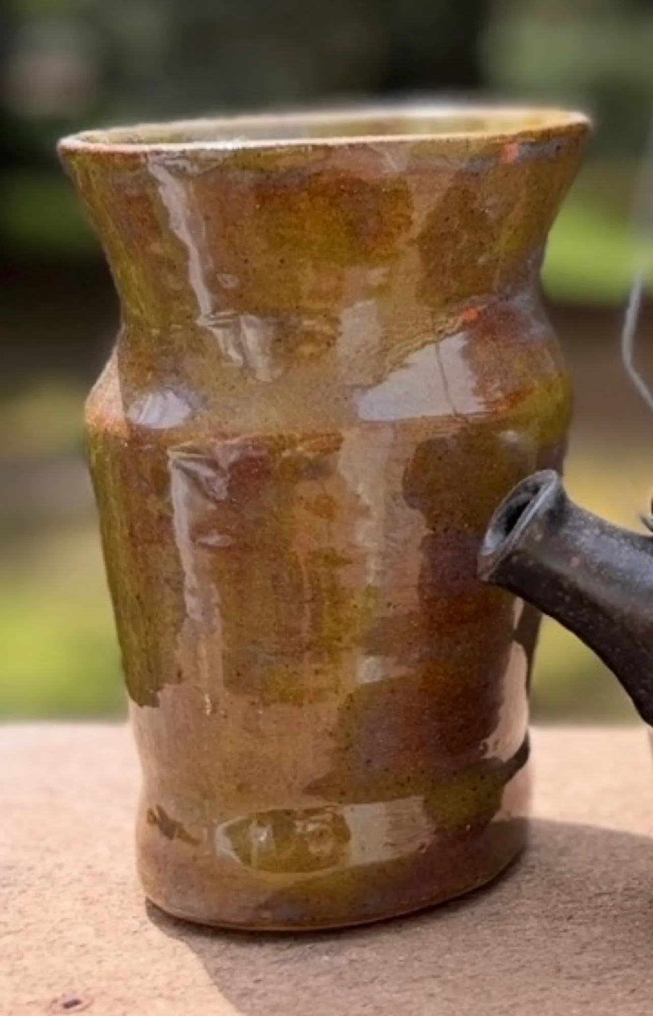 A tall vase with a flared lip in a glossy, mottled olive-green glaze, photographed outdoors.