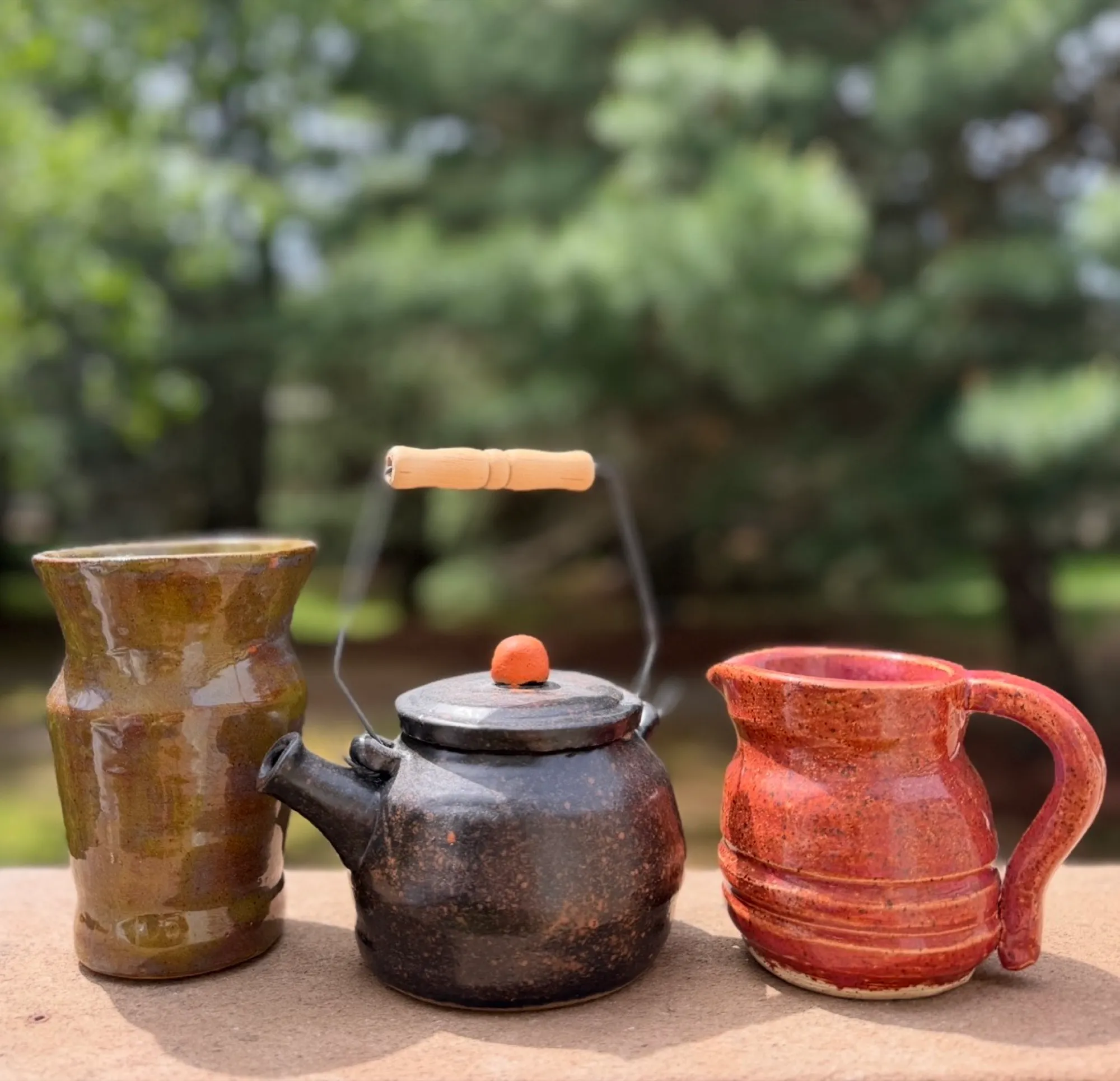 An olive vase, the dark teapot, and a red pitcher lined up on a stone ledge outdoors.