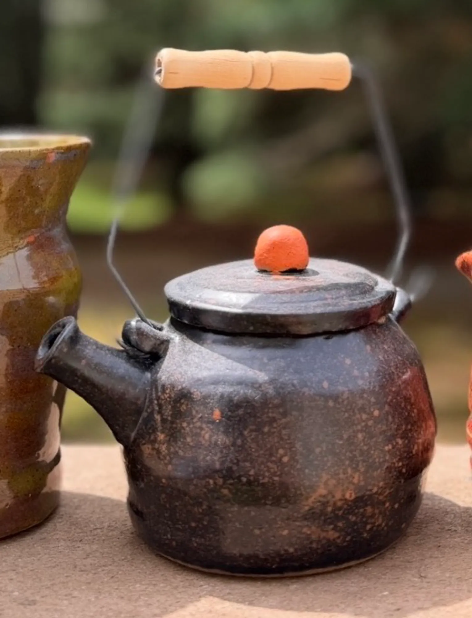 A dark, speckled brown-black teapot with a round terracotta knob on the lid and a curved wire handle with a wooden grip.