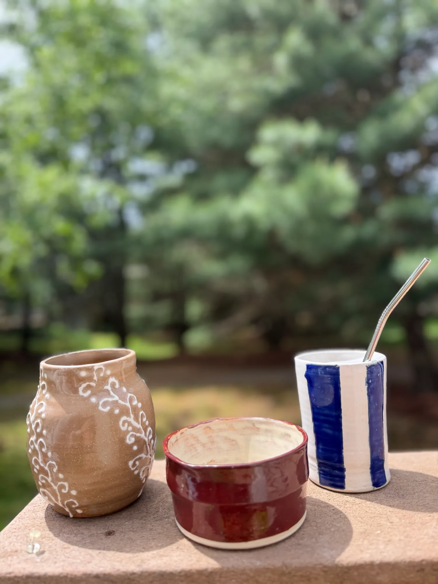 A tan vase with white slip vines, a deep red bowl, and a blue-striped tumbler on a stone ledge in front of pine trees.
